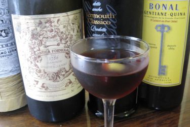 A vermouth cocktail in a cocktail glass with an olive. There are bottles of vermouth in the background.