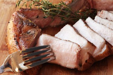 A bone-in cooked pork chop on a cutting board that's been sliced. A fork is in one slice. There is a sprig of fresh thyme next to the pork slices.
