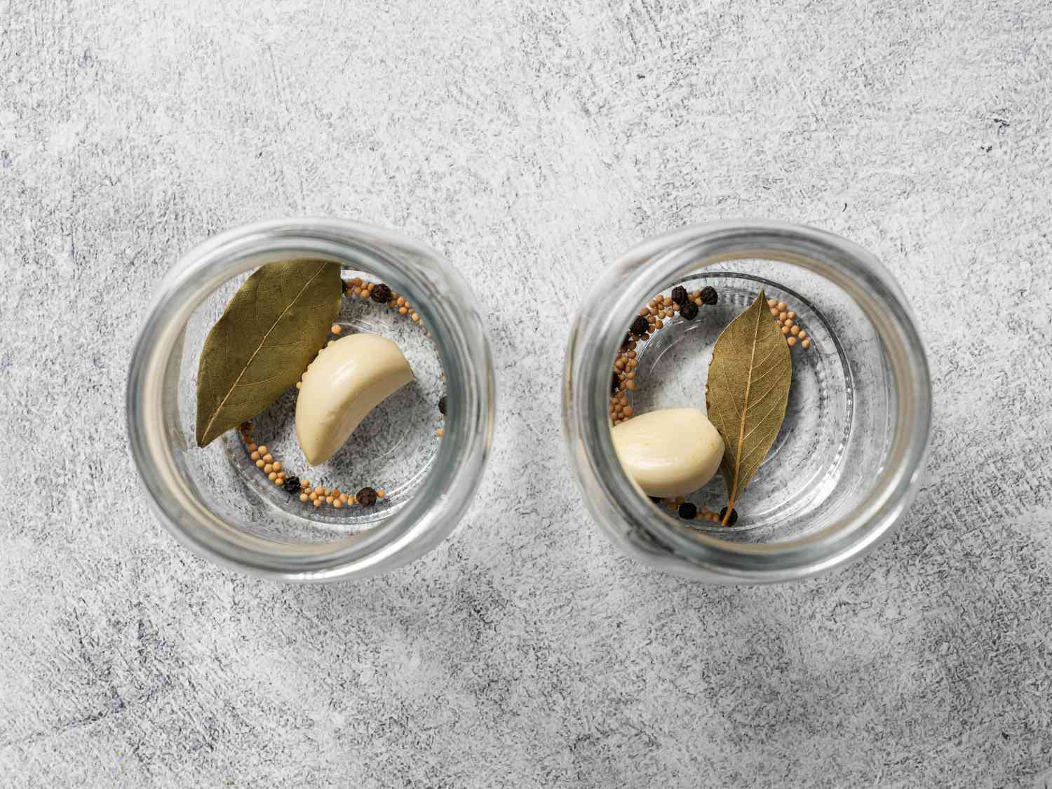 两个玻璃罐并排放置。每个罐子里装着一片月桂叶，一整瓣大蒜，胡椒粒和芥菜籽。