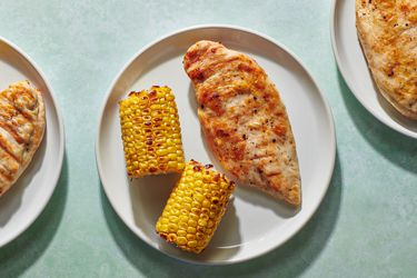 A grilled boneless chicken breast on a white ceramic plate along with two grilled pieces of corn-on-the-cob.