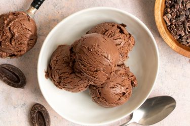 Four scoops of chocolate ice cream in a round white ceramic bowl. Around the periphery of the bowl are a metal spoon, a small wooden bowl holding cacao nibs, two pieces of chocolate, and a scoop holding ice cream.