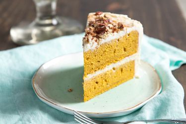 A slice of 2-layer pumpkin layer cake, with a white frosting and chopped pecans on top. The cake is on a small plate with a light blue linen underneath it.