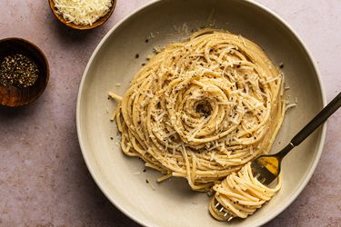 Cacio e pepe on a tan ceramic plate alongside two small bowls, one holding shredded cheese and the other holding freshly ground pepper. There is a metal fork with pasta swirled around it on the plate.