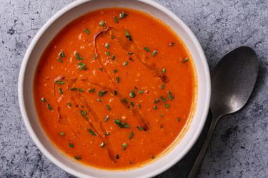 一碗奶油素食番茄汤，在石头背景上，碗的右边有一个勺子。汤的表面淋上橄榄油，撒上切碎的香草。